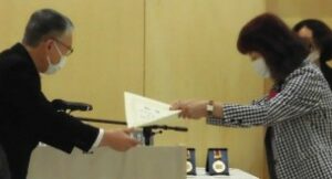 式典でスーツを着た男性から女性に表彰状を手渡している様子の写真