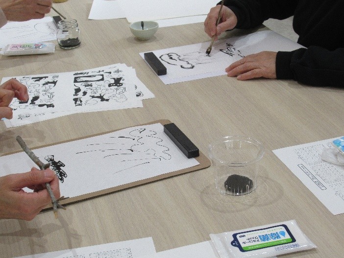 明るい色の木製テーブルを囲み、数人の参加者が小筆や木の枝を筆代わりにして、白い紙に黒い墨で絵や抽象的な線を思い思いに描いている写真