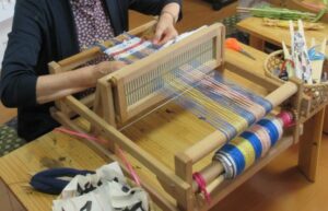 木製の卓上織機で、女性が多色の糸を整えつつ布を織り進めている写真