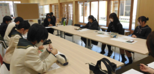 口の字型に配置したテーブルに制服姿の高校生と講師の女性が座り、俳句の作成している様子の写真