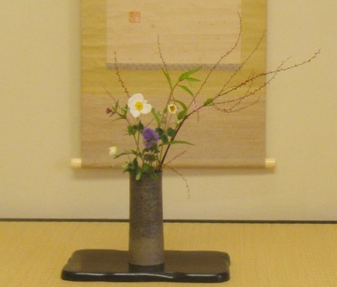 筒形の花器に白や紫の花と細い枝が涼やかに生けられ、掛け軸を背景に飾られている写真