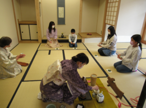 和室の畳の上に参加者が間隔をあけて円座に座り、着物姿の女性が道具を整えて点前を進める中で静かに茶道を体験する様子の写真