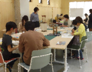 子どもや大人の参加者たちが長机を囲み手元の作業に集中しながら創作活動を行っている様子を写した写真