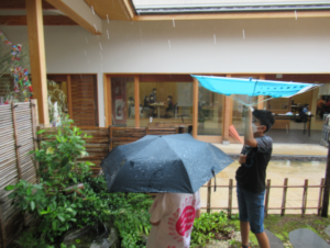軒先の庭で子どもたちが傘を差しながら雨の景色を観察している写真
