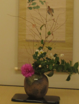 丸い花器に、細い枝や緑の葉と白い小花が付いた枝、濃いピンク色の花が調和して生けられ、掛け軸を背景に飾られている写真