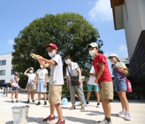 屋外の広場で、バケツの横に竹の水鉄砲を構えた男の子が立ち、複数の子どもたちがその様子を後ろで見守っている様子の写真