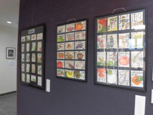 濃紺の壁に、黒い台紙に季節の果物や花などを描いた色彩豊かなたくさんの絵葉書を整然と並べた展示物が3つ、横一列に並んでいる写真