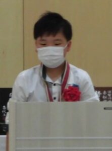 白い演台の前に赤い花飾りを付けた男の子が立っている写真