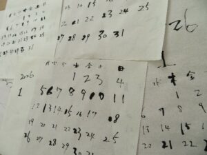 白い和紙の上に、黒い墨と筆を使って「2026」という年号や月、日付、曜日などが手書きで練習するように書き込まれた複数のカレンダー案が、無造作に重なり合うように並べられている様子を写した写真