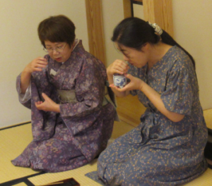 畳に座った女性が、隣に座った和装の女性に作法を習いながら、香りをかいでいる様子の写真