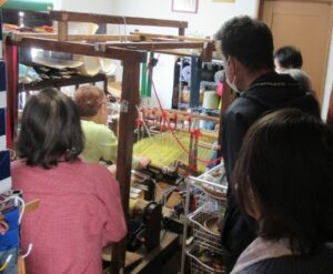 木製の機織り機で作業する様子を周囲の人々が間近で見学している様子の写真