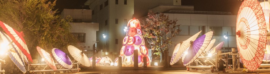 夜の街並みに色鮮やかな和傘が灯りを放ちながら左右に並び、中央には多数の和傘で形作られた塔が輝く情景の写真