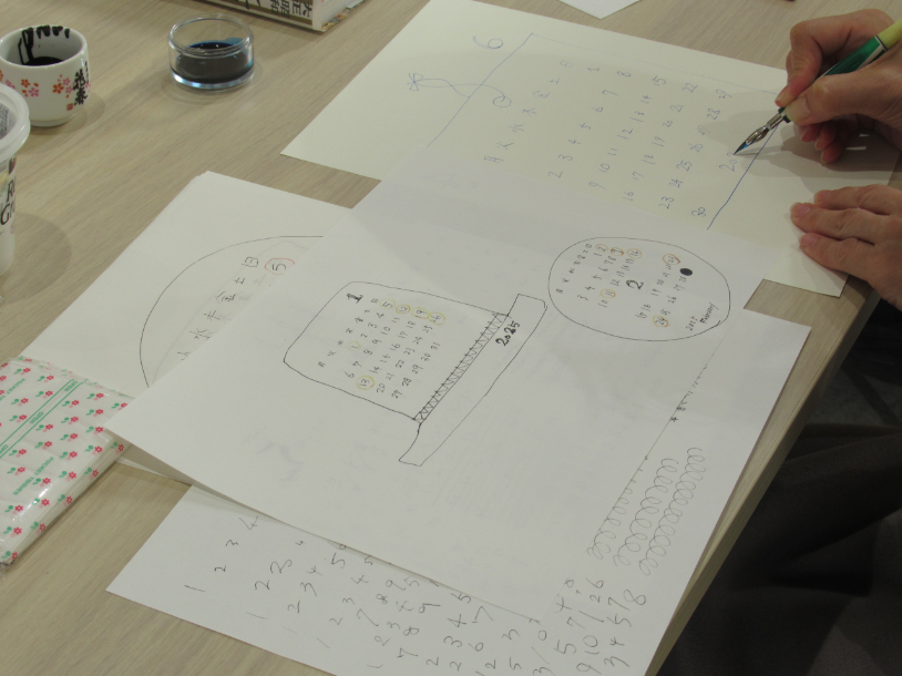 手書きの帽子をモチーフにしたカレンダーや数字の練習が描かれた複数の紙が広げられた横で、つけペンを持ってカレンダーの数字を丁寧に書き込んでいる手元の写真
