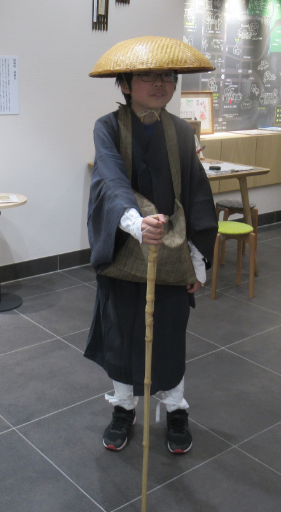 濃い灰色の旅装束に編み笠をかぶり杖を携えた子どもが室内で松尾芭蕉の姿を再現して立っている写真