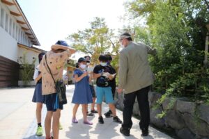 屋外の植え込みの前で複数の子どもたちが男性の周りに集まり、植物などを観察しながらメモを取っている様子の写真