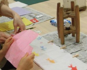 色とりどりの金魚が配置された白い和紙と糸巻きを置いたテーブルの前に座わり、ピンク色の色紙を持っている子どもの手元の写真