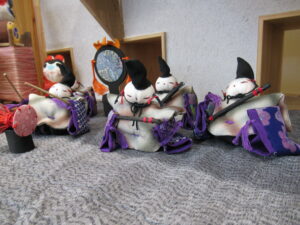 紫の装飾が付いた白の衣装をまとい、烏帽子をかぶった小さな布製人形が太鼓や笛を手に並んで演奏するように配置された展示の写真