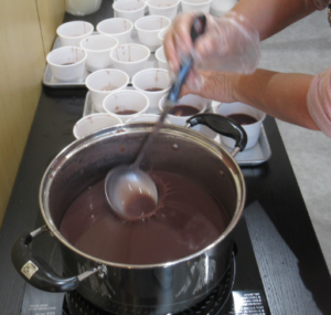 大きな鍋に入ったたっぷりのおしるこを、お玉ですくい、トレイに並べられた多数の白いカップへ取り分けている写真