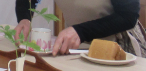 一輪挿しが飾られた卓上に、ケーキが乗った小皿が置かれ、その隣に、ピンク色の花が描かれたティーカップが乗ったソーサーを丁寧に置いている様子の写真