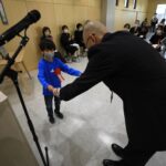 木目調の明るい室内で、青い服に赤いリボンをつけたマスク姿の男の子が、スーツ姿の男性から白い賞状を両手で受け取っている表彰式の写真