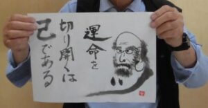 達磨の絵と「運命を切り開くは己である」の文字が墨で書かれた半紙が両手で掲げられている様子の写真