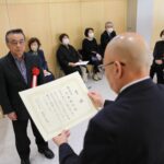 木目調の明るい室内で、手前に立つ男性が「賞状」と書かれた証書を両手に持っており、赤いリボンを胸に付けたスーツ姿の男性に授与している様子を捉えた写真