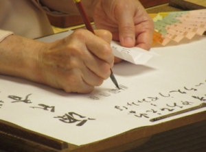 右手で細い筆を使い、左手に持つ白い紙を見ながら、用紙に文字を丁寧に書き記している写真
