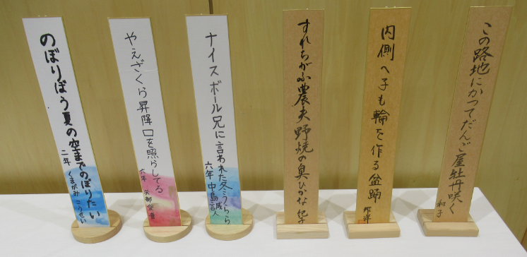 木目調の背景を背に、俳句や短歌が縦書きで記された白や茶色の短冊6枚が木製の台座に立てられて一列に並んでおり、子供から大人まで多様な作者による作品が展示されている写真