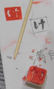 ひらがなで「け」と彫られた小さな四角の消しゴム印鑑が、印鑑のデザインが書かれ、試し押しがされた紙の上に置かれている写真