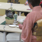 薄ピンク色の着物の女性、茶釜や茶器が準備されたテーブルの前に立っている後ろ姿の写真