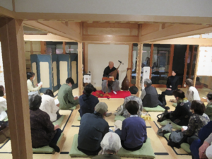 和室で赤い毛氈の上に座る奏者が伝統的な弦楽器である薩摩琵琶を演奏しており、その前で大勢の観客が座布団に座って静かに聴き入っている写真