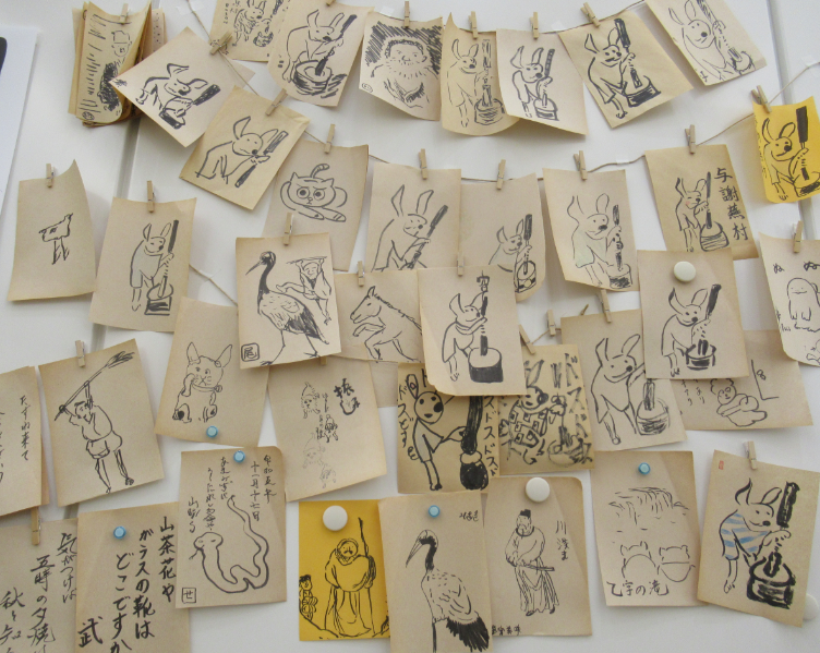 餅をつくウサギや人物などが黒で描かれた、たくさんの正方形の小さな紙が、重なりながら壁一面にクリップやマグネットで留められている写真