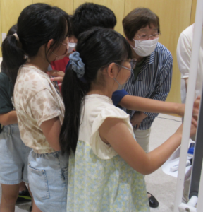 教室で数人の子どもや大人がホワイトボードの前に集まり、掲示内容を熱心に確認している写真