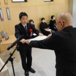 木目調の明るい室内で、黒いスーツに赤いリボンをつけたマスク姿の男性が、スーツ姿の男性から白い賞状を両手で受け取っている表彰式の写真