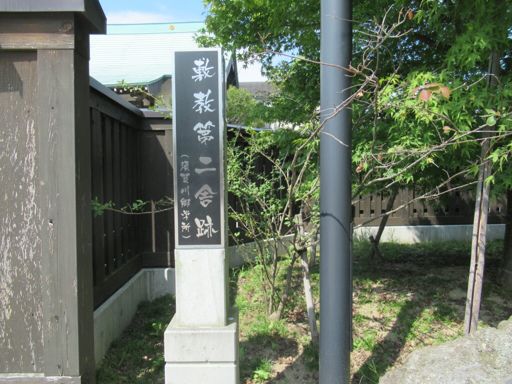こげ茶色の木製の囲いの内側に、木々に囲まれて、黒い背景に「敷教第二舎跡（須賀川郷学所）」と白抜きの文字で刻まれた標柱が立っている写真