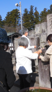 青空の下、たくさん並んだ古い石碑に数名の男女が花を手向けて並んでいる様子の写真