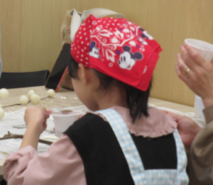 赤いバンダナを巻いた女の子が、テーブルの上に置かれた白いカップに入ったおしるこを食べている様子を後ろから捉えた写真