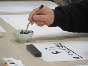 テーブルで、参加者が木の枝を筆代わりにして小さな器の墨を浸し、白い和紙に「日月火水木金土」という曜日などの文字を独特の筆致で丁寧に書き進めている様子を近くから捉えた写真