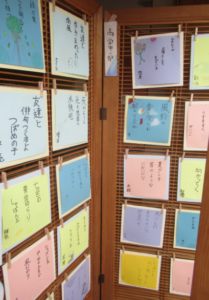 中央に「高学年の部」の短冊が貼られた格子状の木製パネルに、俳句やイラストが手書された様々な色の色紙がたくさん並んでいる展示の様子の写真