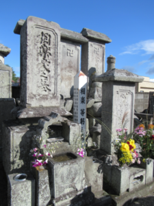 青空の下で風化した複数の石造墓碑が立ち並び色とりどりの供花が手向けられて静かに佇む墓所の写真