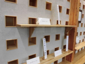 格子状に四角い穴が並ぶ白い壁に設置された細い棚に、短冊形の作品が点在して配置された落ち着いた展示空間の写真