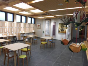 テーブルと椅子のセットが複数置かれた、オープンギャラリーの左側の壁一面に、俳句作品が貼られた木のパネルが設置されている様子の写真