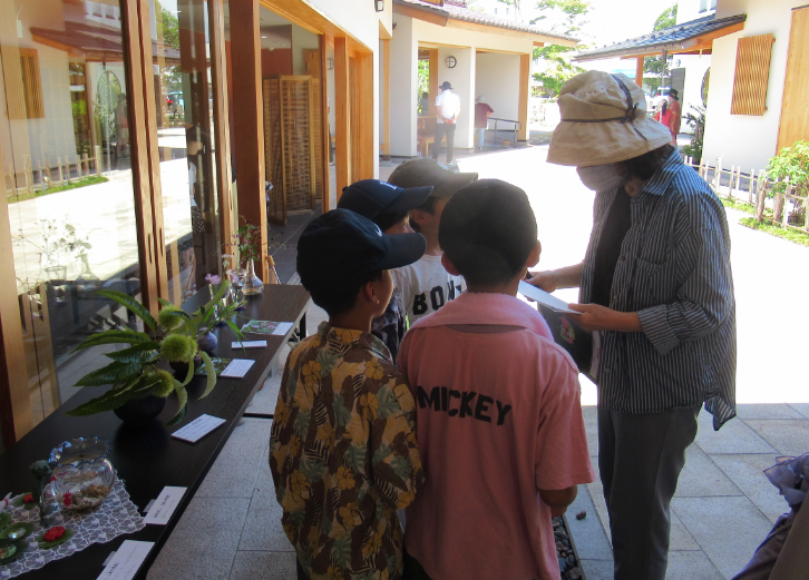 屋外の回廊で帽子をかぶった子どもたちが女性から説明を受けつつ生け花作品を見学する写真