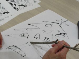 テーブルの上で、参加者が細い木の枝を筆の代わりにして、白い和紙に黒い墨でクリスマスツリーのイラストを描いている写真