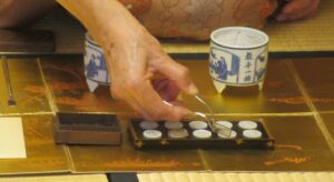 落ち着いた和室の畳の上で金屏風のような敷物を前に、着物姿の人物が銀色の火箸を使い、並べられた小さな白い香道具を丁寧に扱う香道の繊細な手元の様子を捉えた写真