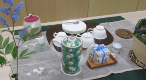 テーブルに敷かれたクロスの上に、花柄のポットやカップ、ソーサーなどが並び、青い小花が鈴なりに咲いた枝の一輪挿しが飾られている写真