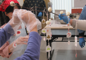 三角巾やエプロンをつけた参加者が、木の枝に白い団子や吊るし飾りを飾り付けている様子の写真