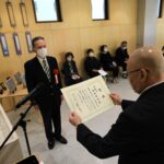 木目調の明るい室内で、黒いスーツ姿の男性が賞状を両手で掲げ、その正面に立つマスク姿の男性に授与している様子を捉えた写真