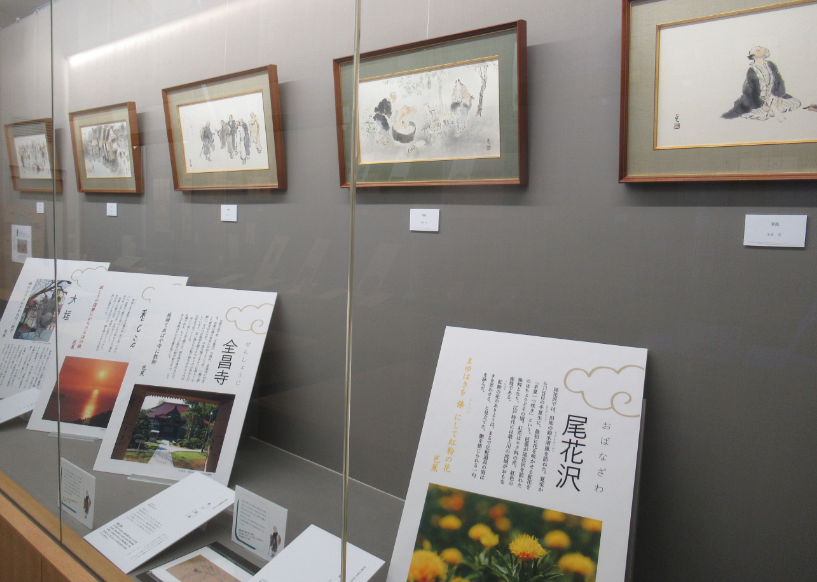 ガラスの展示ケース内に複数の日本画と、関連した情景を解説するパネルが整然と並ぶ展示の様子の写真