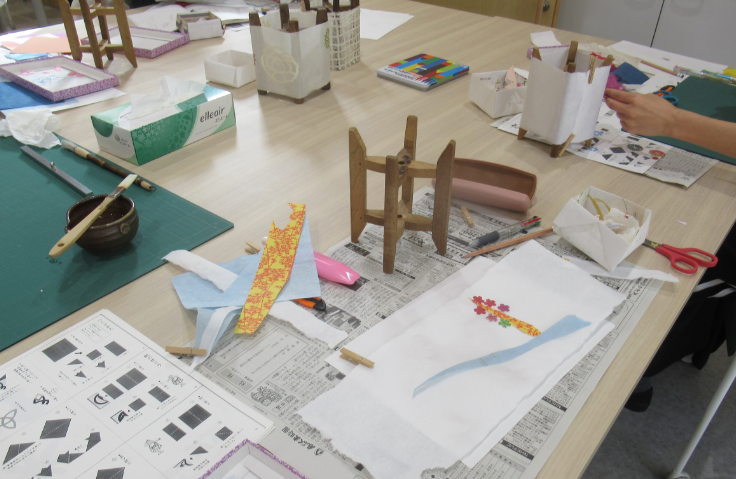 作業台の上に糸巻きや花などを貼った和紙、ハサミやノリが置かれている作業中の様子の写真